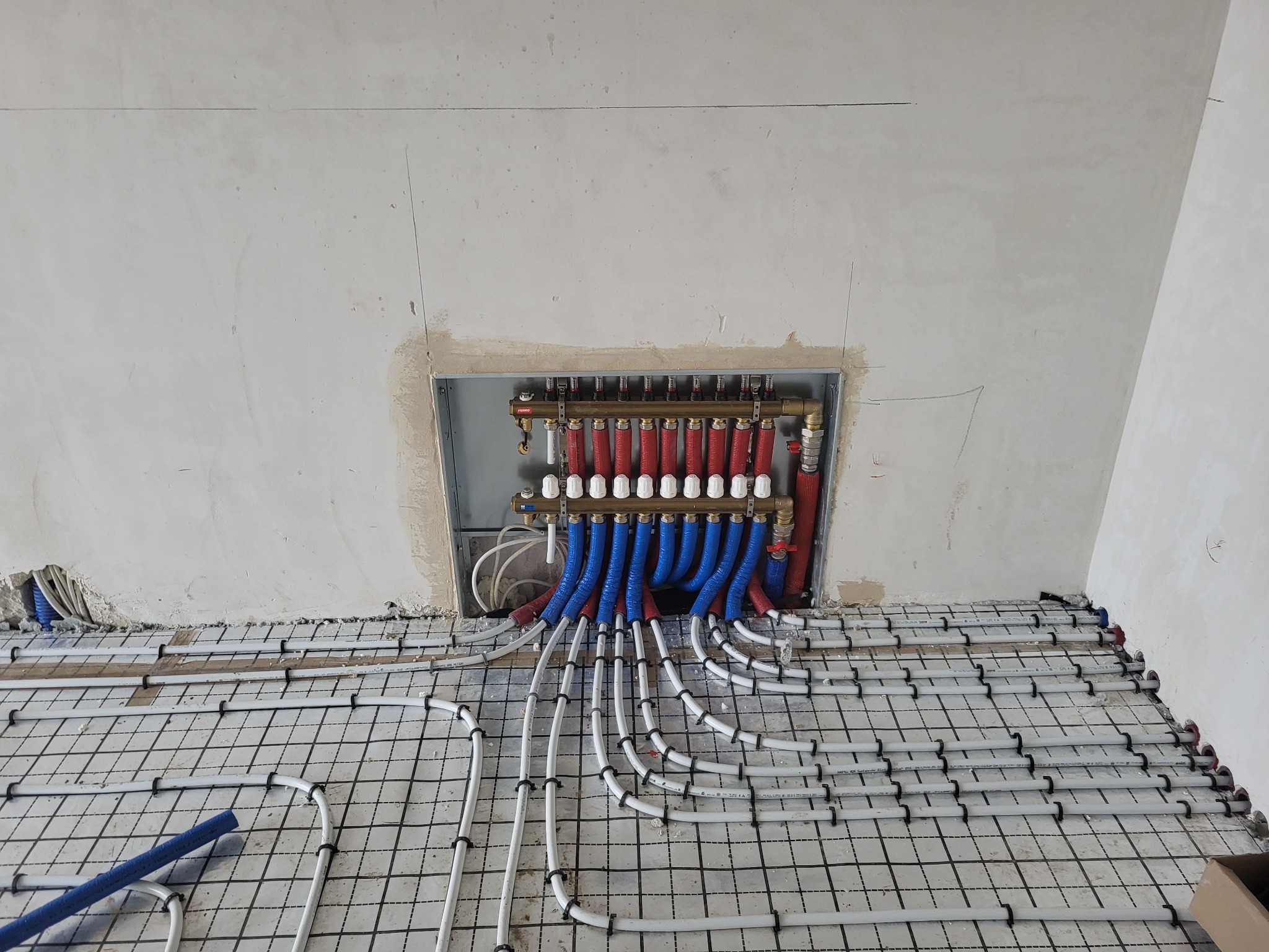 Rozdzielacz ogrzewania podłogowego z niebieskimi i czerwonymi rurami w Podbrzeziu Dolnym, widok na siatkę montażową i surową ścianę.