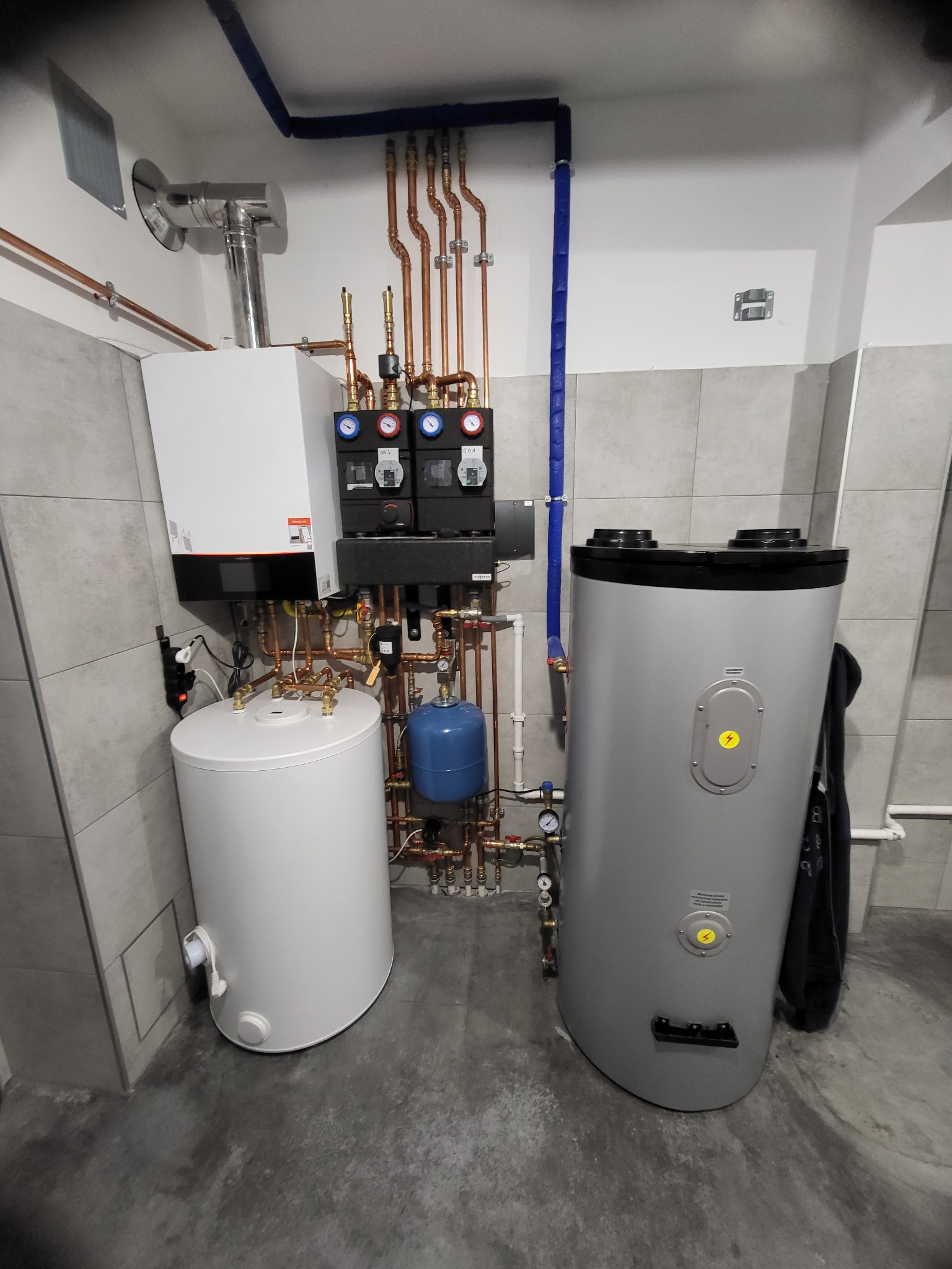 Kotłownia z nowoczesnym kotłem grzewczym, zasobnikiem ciepłej wody, zbiornikiem wyrównawczym i bogatą instalacją z rur miedzianych, zabezpieczonych niebieską izolacją.