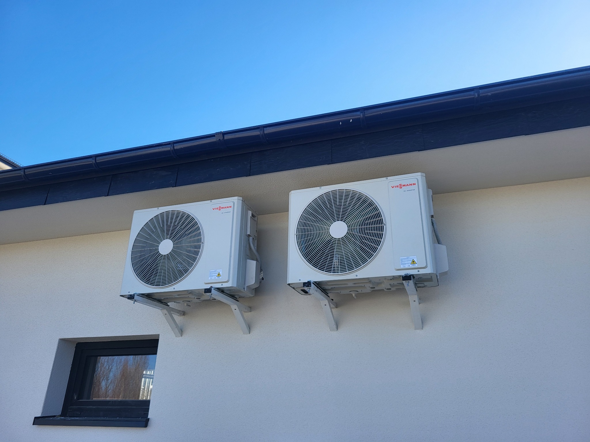 Dwa identyczne, białe klimatyzatory Viessmann zamontowane na wspornikach na elewacji budynku z czarnym oknem i rynną na tle błękitnego nieba.