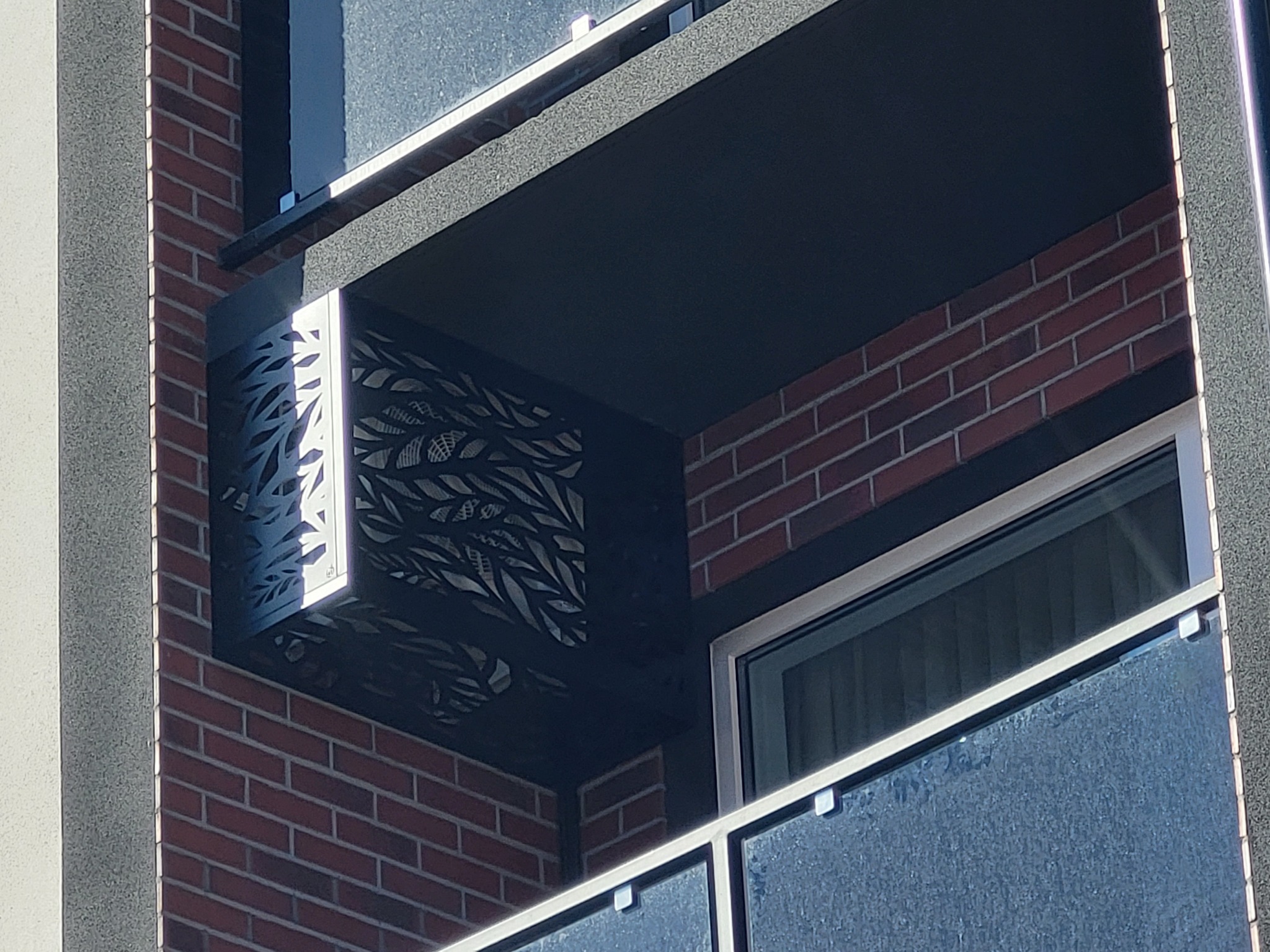 Fragment balkonu z dekoracyjną, ażurową osłoną z motywem liści, ceglana elewacja budynku w tle, widoczne okno i fragment szklanej balustrady.