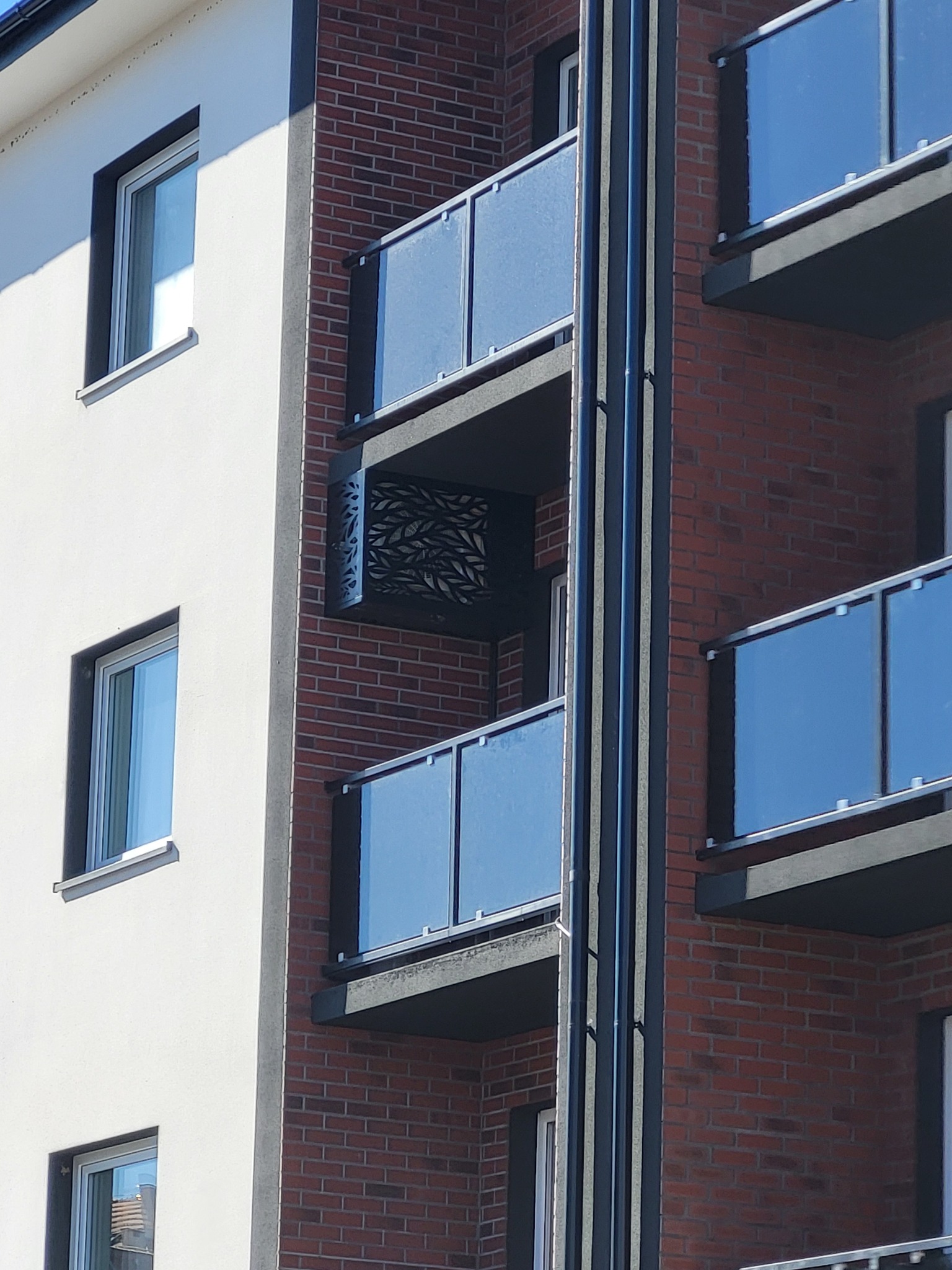 Fragment fasady budynku z balkonami z szklanymi balustradami i dekoracyjnymi panelami ażurowymi, widoczne pionowe rury instalacyjne.