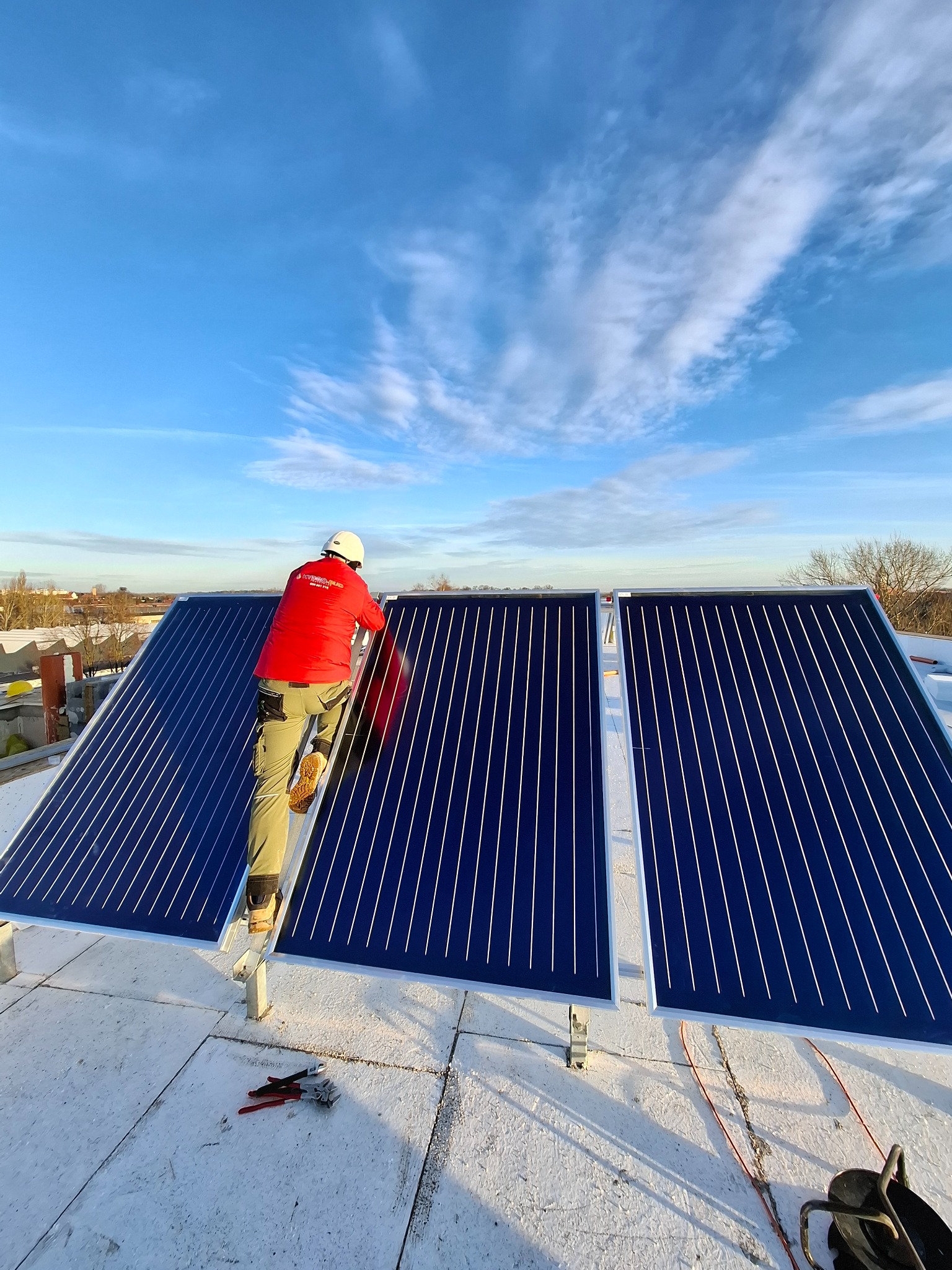 Instalacja solarna na dachu: monter w czerwonej kurtce na drabinie, panele z niebieskimi liniami, narzędzia obok. Błękitne niebo w tle.