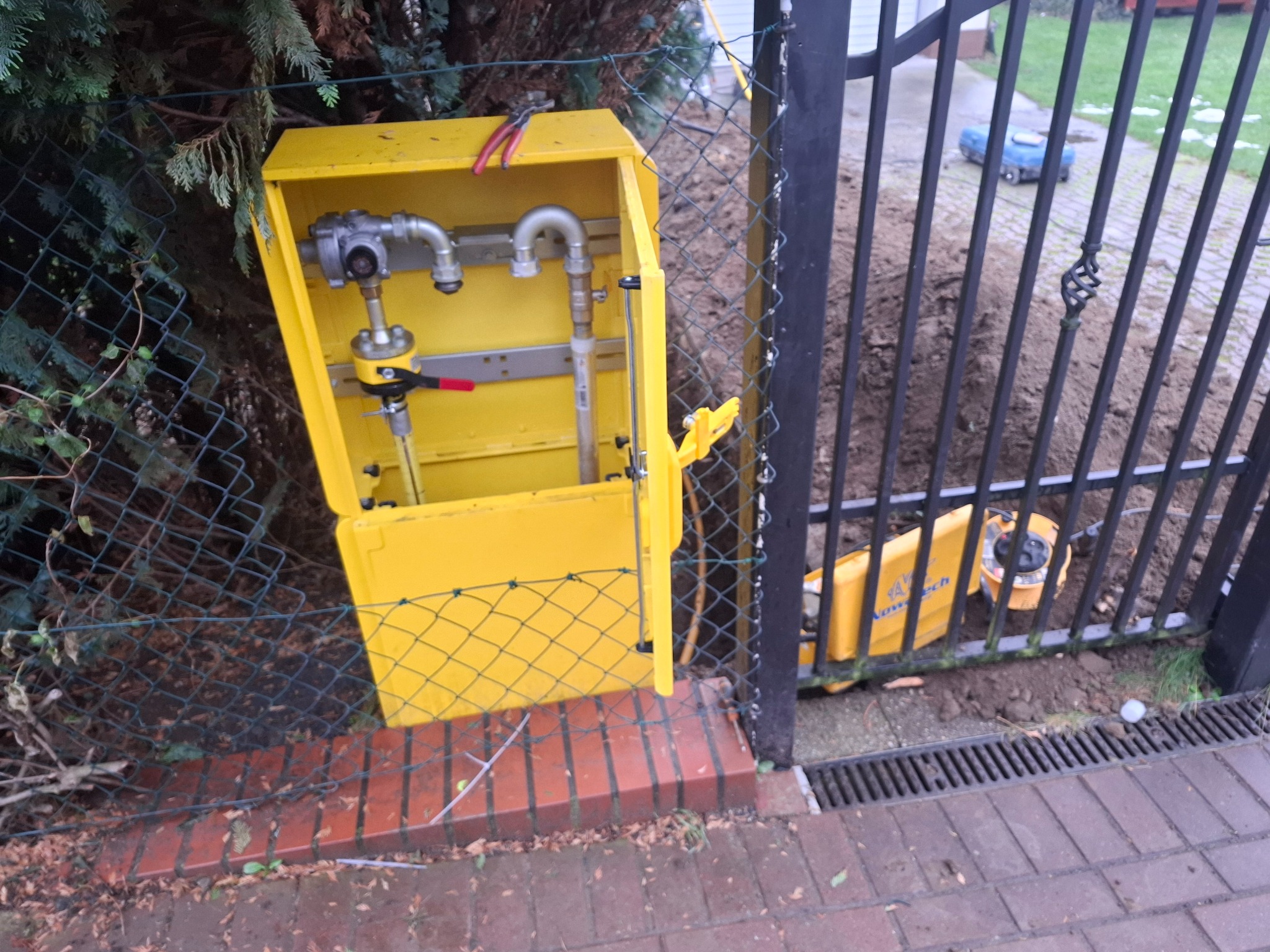 Żółta skrzynka gazowa z otwartymi drzwiczkami, widoczne elementy instalacji gazowej, obok ogrodzenie i wykop, sugerujące prace przy przyłączu gazowym.