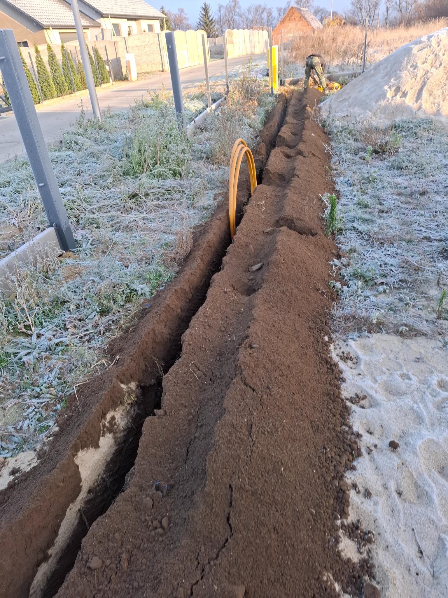 Wykopany rów z żółtymi rurami instalacji gazowej w ziemi, widoczny pracownik w oddali, lekko oszroniona trawa, w tle domy.