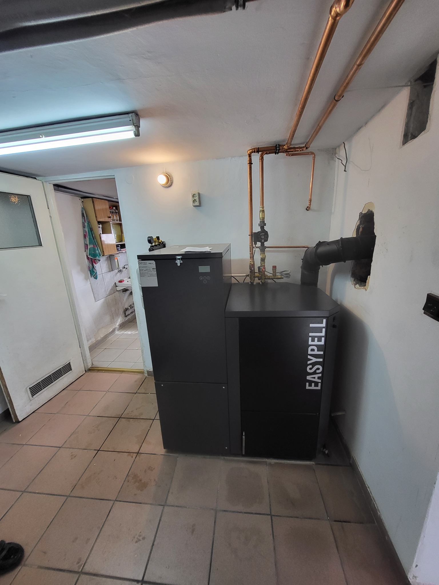 Czarny piec EasyPell w piwnicy z widocznymi miedzianymi rurami instalacji grzewczej. W tle otwarte drzwi do pomieszczenia z kafelkami i umywalką.