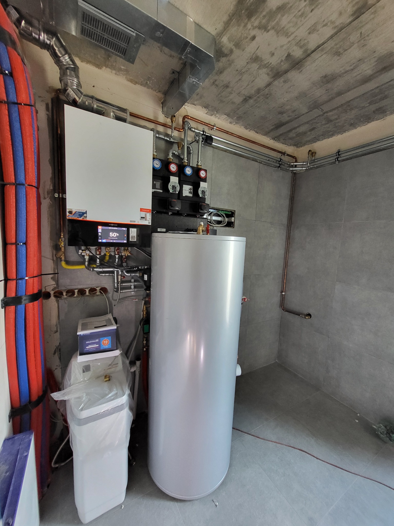 Nowoczesna kotłownia z piecem gazowym, zasobnikiem ciepłej wody i zmiękczaczem wody. Widoczne rury instalacji grzewczej i wodnej. Szara płytka na ścianach i betonowy sufit.