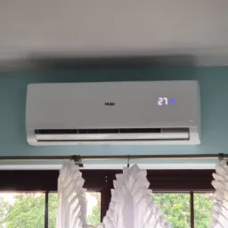 Biała klimatyzacja Haier zamontowana na błękitnej ścianie nad oknem z białymi, dekoracyjnymi zasłonami, widok na zieleń z zewnątrz, wyświetlacz pokazuje temperaturę 27 stopni.