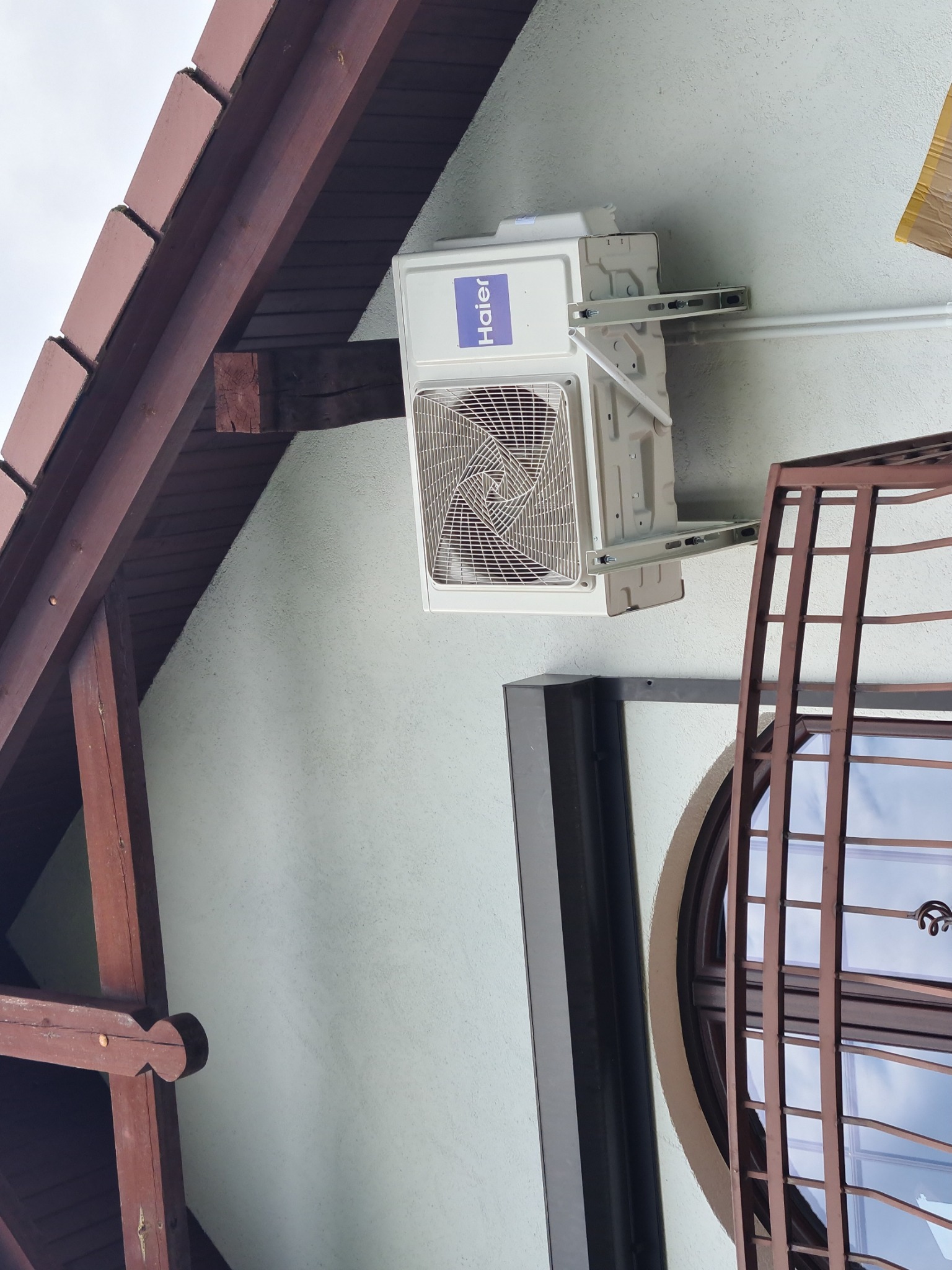 Biała jednostka zewnętrzna klimatyzatora Haier zamontowana na ścianie domu z drewnianą konstrukcją dachu i okrągłym oknem z ozdobną kratą.
