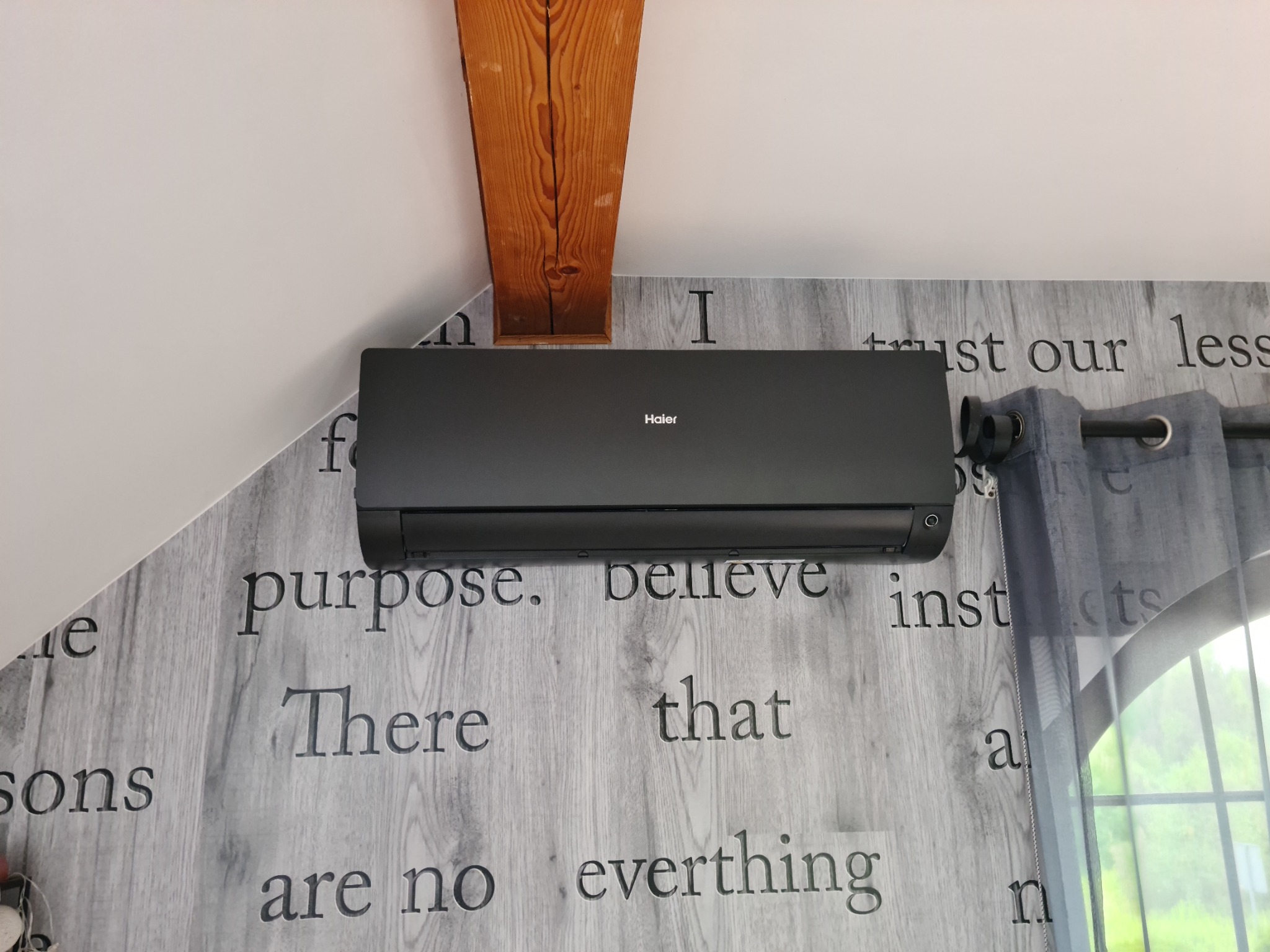 Czarna jednostka klimatyzacji Haier zamontowana na ścianie z tapetą imitującą drewno z wygrawerowanymi słowami, widoczny fragment drewnianej belki stropowej i zasłony w tle.