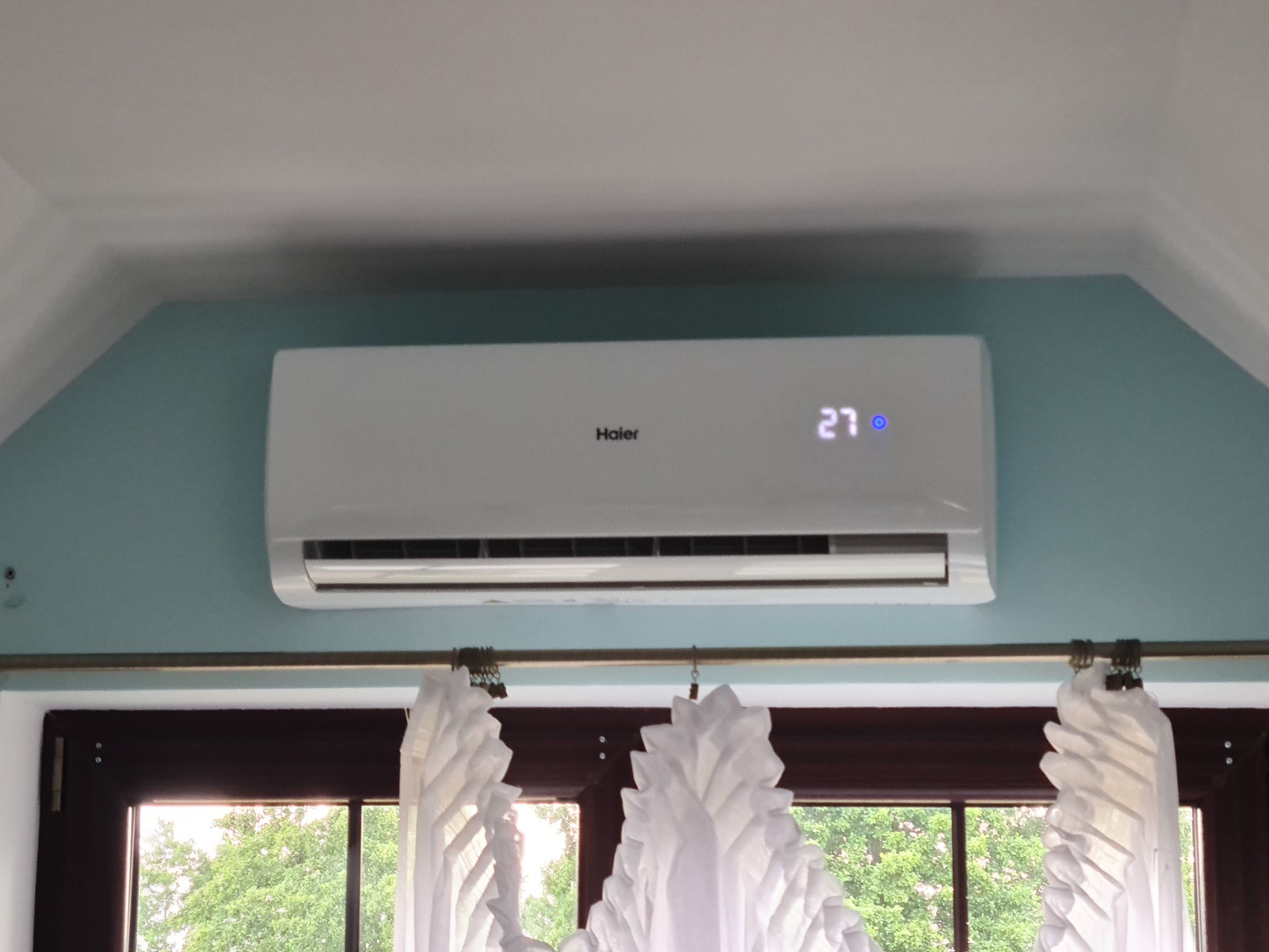 Biała klimatyzacja Haier zamontowana na błękitnej ścianie nad oknem z białymi, dekoracyjnymi zasłonami, widok na zieleń z zewnątrz, wyświetlacz pokazuje temperaturę 27 stopni.