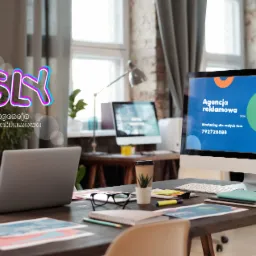 Biuro agencji reklamowej z widocznym logotypem SLY, laptopem, monitorem wyświetlającym wykres kołowy z danymi marketingowymi oraz biurkiem z okularami i notatnikiem.