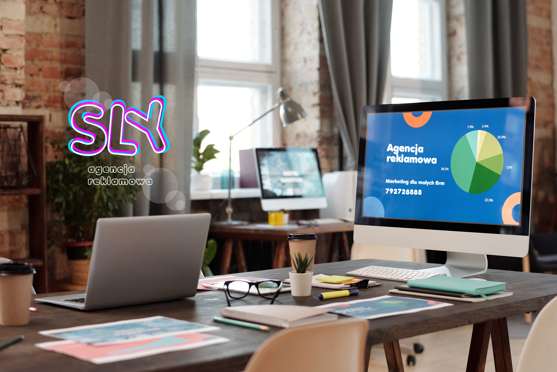 Biuro agencji reklamowej z widocznym logotypem SLY, laptopem, monitorem wyświetlającym wykres kołowy z danymi marketingowymi oraz biurkiem z okularami i notatnikiem.