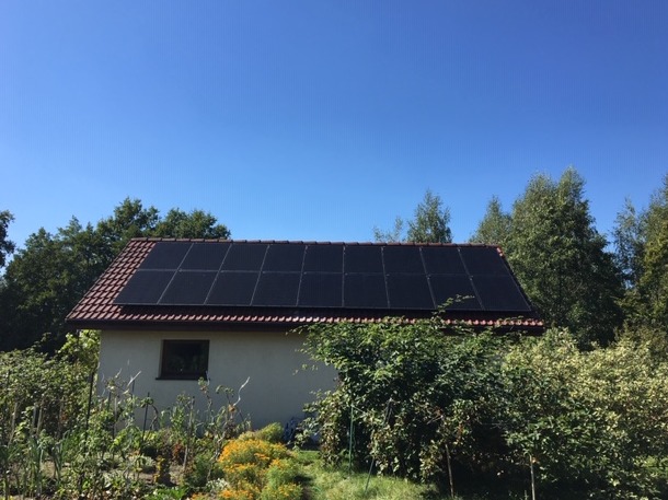 Dom jednorodzinny z panelami słonecznymi na dachu, otoczony zielenią, widok z poziomu ogrodu, błękitne niebo w tle.