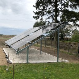 Wojciech Pawluczuk - Instalacja paneli słonecznych na konstrukcji wsporczej, podsypana białym żwirem, na tle zielonej trawy i pochmurnego nieba.