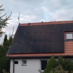 Wojciech Pawluczuk - Instalacja paneli słonecznych na dachu domu z czerwoną dachówką, widoczna antena i okno dachowe, otoczenie drzewa iglaste.