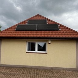 Wojciech Pawluczuk - Niewielki budynek z żółtą elewacją i czerwoną dachówką, na którym zamontowano cztery czarne panele słoneczne, widoczne białe okno i oświetlenie zewnętrzne, pochmurne niebo w tle.