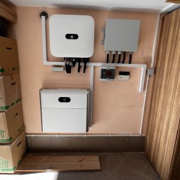 Wojciech Pawluczuk - Zamontowany inwerter i magazyn energii firmy Huawei na pomarańczowej ścianie, widoczne kable i zabezpieczenia, na podłodze leżą deski.
