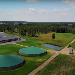 Wojciech Pawluczuk - Panoramiczny widok z lotu ptaka na biogazownię z dwoma zielonymi zbiornikami, budynkiem gospodarczym i niewielką instalacją paneli słonecznych na tle zielonych pól i lasów, w słoneczny dzień.
