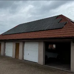 Panele słoneczne na dachu garażu z trzema bramami garażowymi, widoczne auto w jednym z garaży, pochmurne niebo