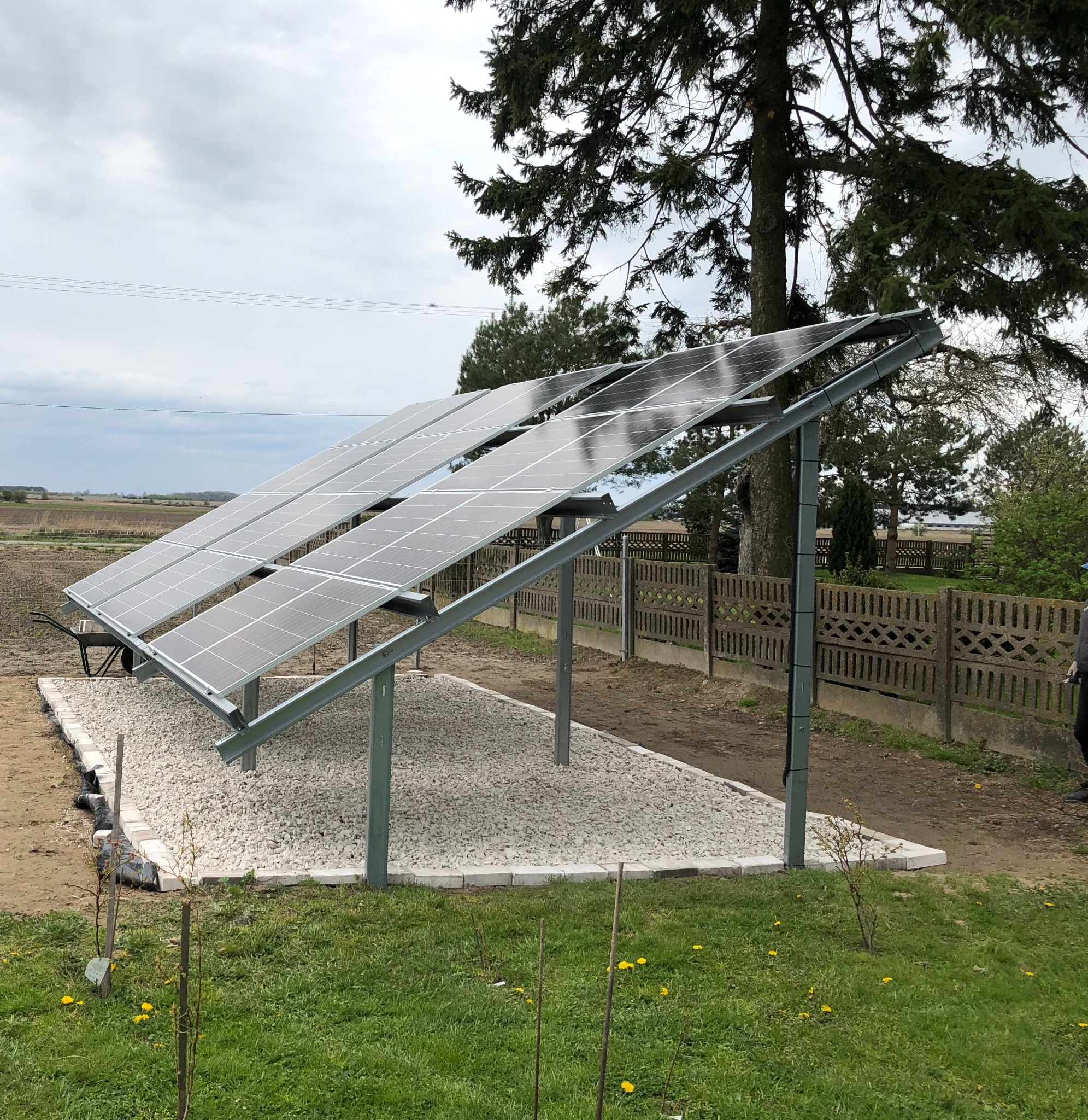 Instalacja paneli słonecznych na konstrukcji wsporczej, podsypana białym żwirem, na tle zielonej trawy i pochmurnego nieba.