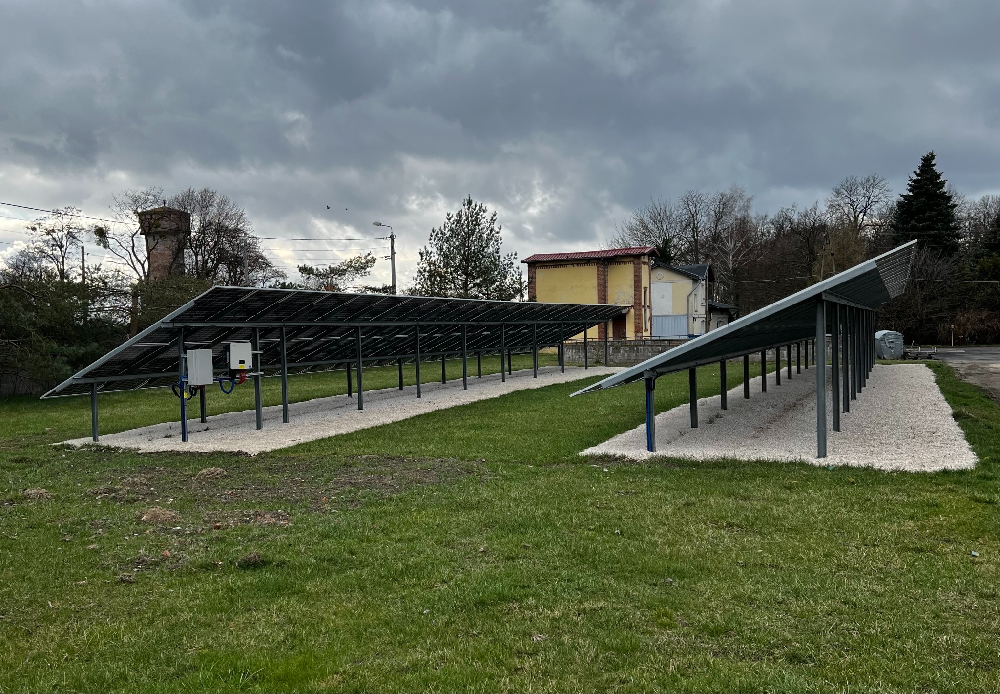 Dwa rzędy paneli słonecznych na metalowych stelażach, ułożone na białym żwirze, z budynkiem w tle pod pochmurnym niebem.