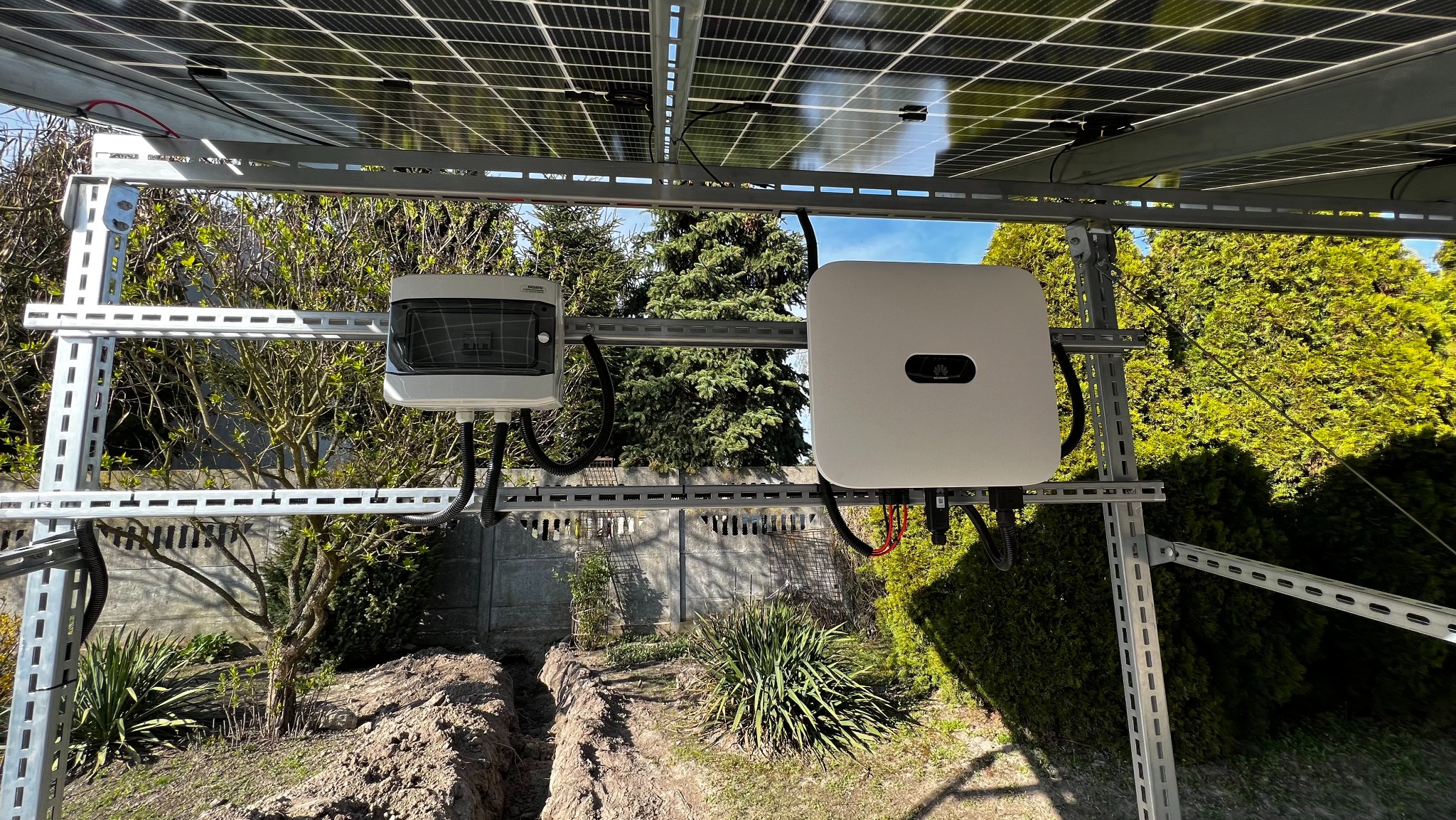 Widok z dołu na zainstalowane panele słoneczne, biały falownik Huawei i skrzynkę przyłączeniową na metalowej konstrukcji montażowej, z widocznym wykopem w ziemi i zielenią w tle.