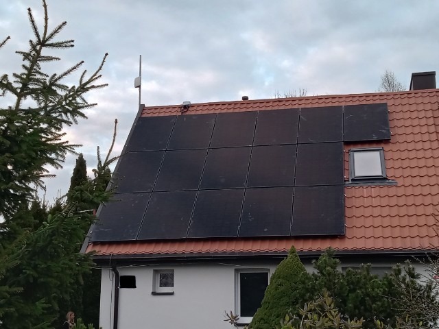 Instalacja paneli słonecznych na dachu domu z czerwoną dachówką, widoczna antena i okno dachowe, otoczenie drzewa iglaste.