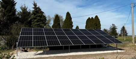 Instalacja paneli słonecznych w Warszawie, umieszczona na metalowej konstrukcji z podłożem wysypanym białym żwirem, widoczna na tle zielonych drzew i błękitnego nieba.