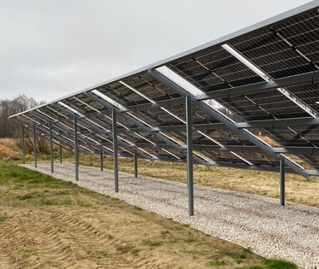 Widok od spodu na konstrukcję wsporczą paneli fotowoltaicznych, z widocznymi rzędami paneli, stalowymi słupkami i wysypanym żwirem pod konstrukcją na tle jesiennego krajobrazu.