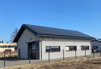 Biały budynek z garażem i panelami słonecznymi na dachu, widok zza ogrodzenia, błękitne niebo w tle.