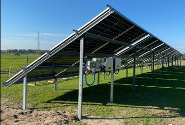 Konstrukcja nośna paneli słonecznych z widocznymi inwerterami i okablowaniem, zamontowana na zielonej trawie w słoneczny dzień.
