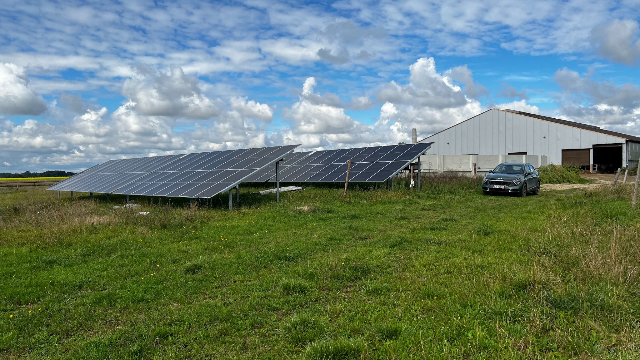 Instalacja paneli słonecznych na łące, z widocznym budynkiem gospodarczym w tle i zaparkowanym samochodem osobowym.