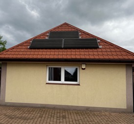 Niewielki budynek z żółtą elewacją i czerwoną dachówką, na którym zamontowano cztery czarne panele słoneczne, widoczne białe okno i oświetlenie zewnętrzne, pochmurne niebo w tle.