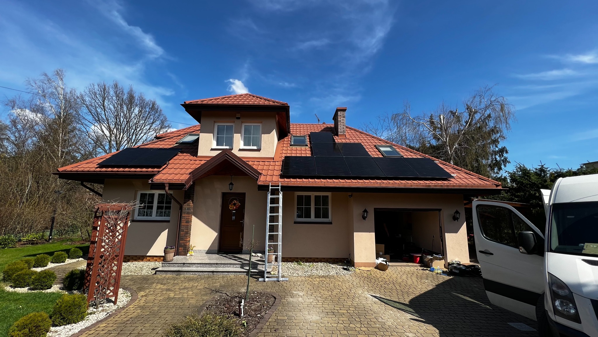 Dom jednorodzinny z czerwoną dachówką, na której zamontowane są czarne panele słoneczne. Przy ścianie domu stoi aluminiowa drabina. Częściowo widoczny biały samochód dostawczy z otwartymi drzwiami.