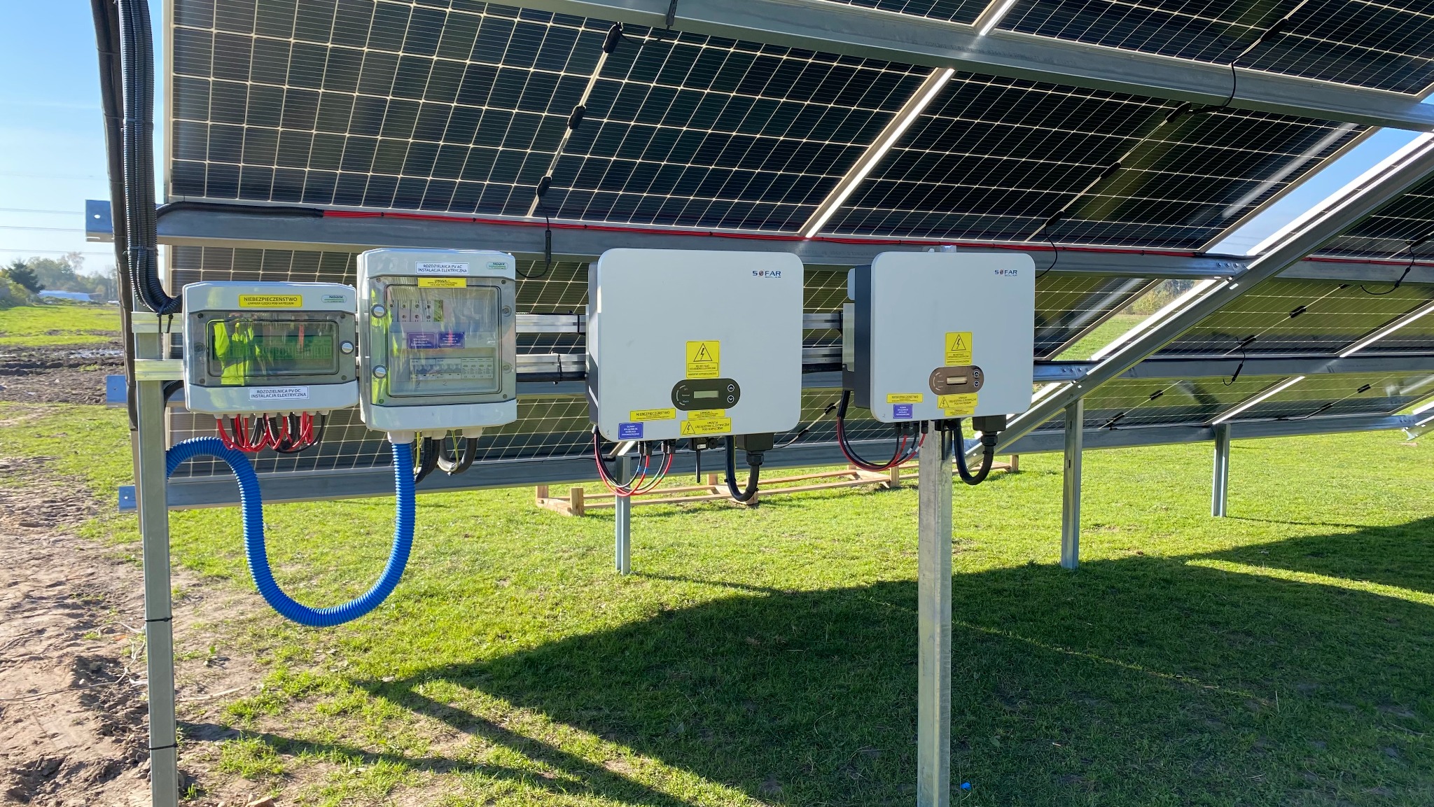 Instalacja fotowoltaiczna: widok od dołu na panele słoneczne, dwa inwertery marki Sofar i skrzynki zabezpieczeń na metalowych konstrukcjach, na tle zielonej trawy i błękitnego nieba.