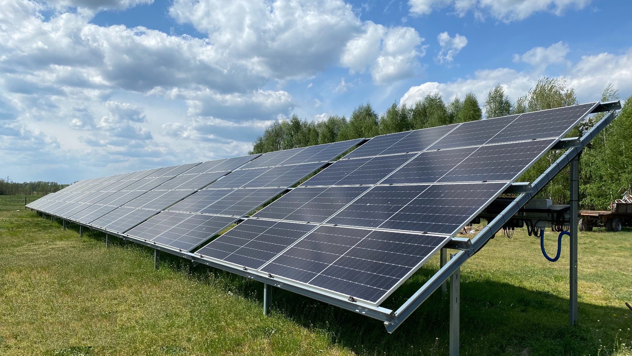 Długi rząd paneli słonecznych na metalowej konstrukcji, zainstalowany na zielonej łące pod błękitnym niebem z białymi chmurami. W tle widoczny las.