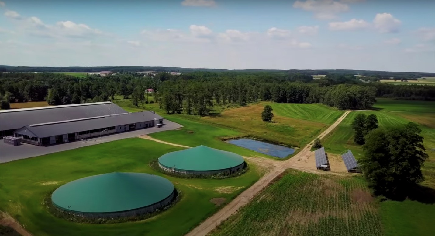 Panoramiczny widok z lotu ptaka na biogazownię z dwoma zielonymi zbiornikami, budynkiem gospodarczym i niewielką instalacją paneli słonecznych na tle zielonych pól i lasów, w słoneczny dzień.
