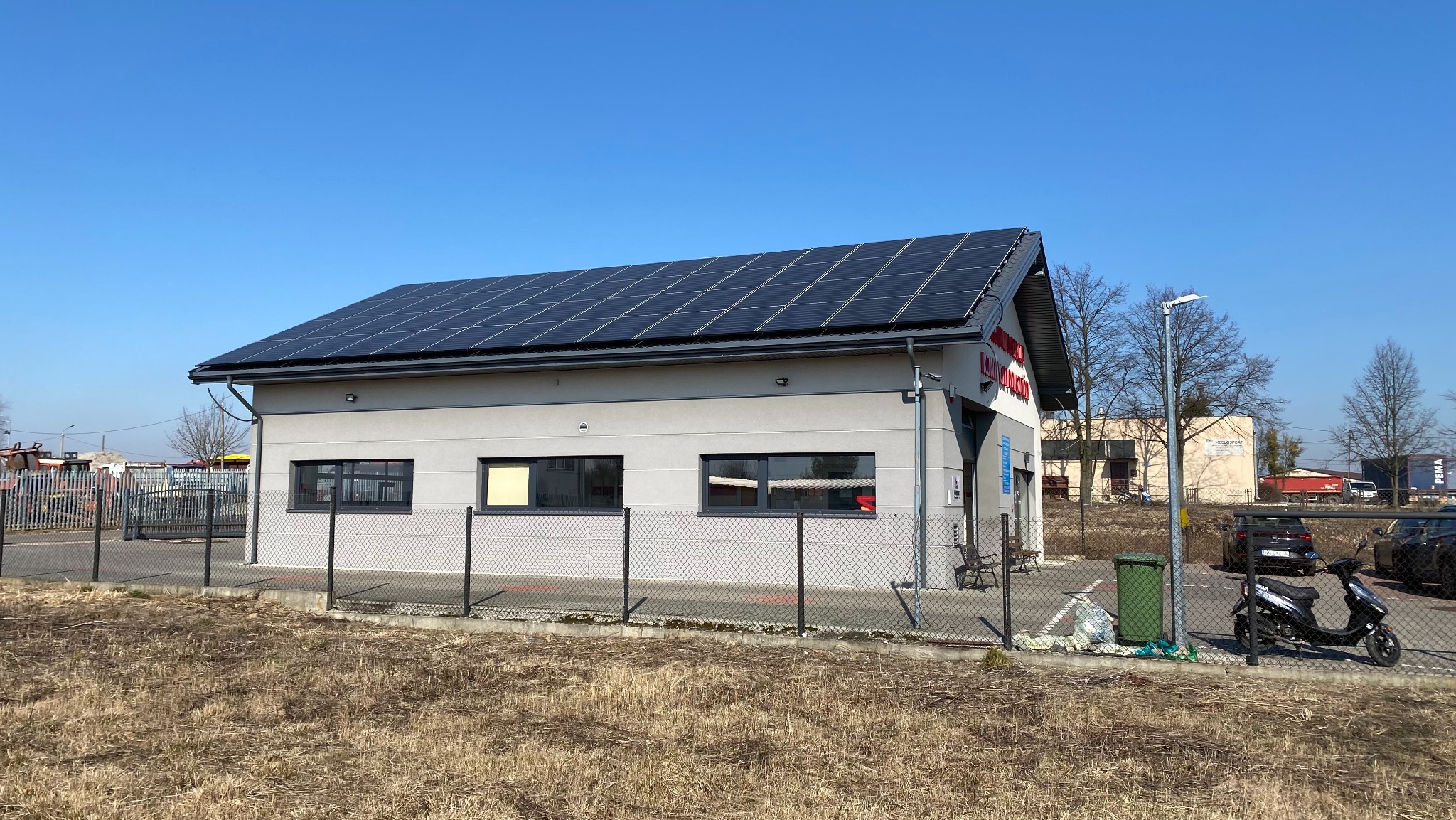 Szary budynek z panelami słonecznymi na dachu, ogrodzony siatką, z motocyklem zaparkowanym przed nim i zielonym koszem na śmieci obok.