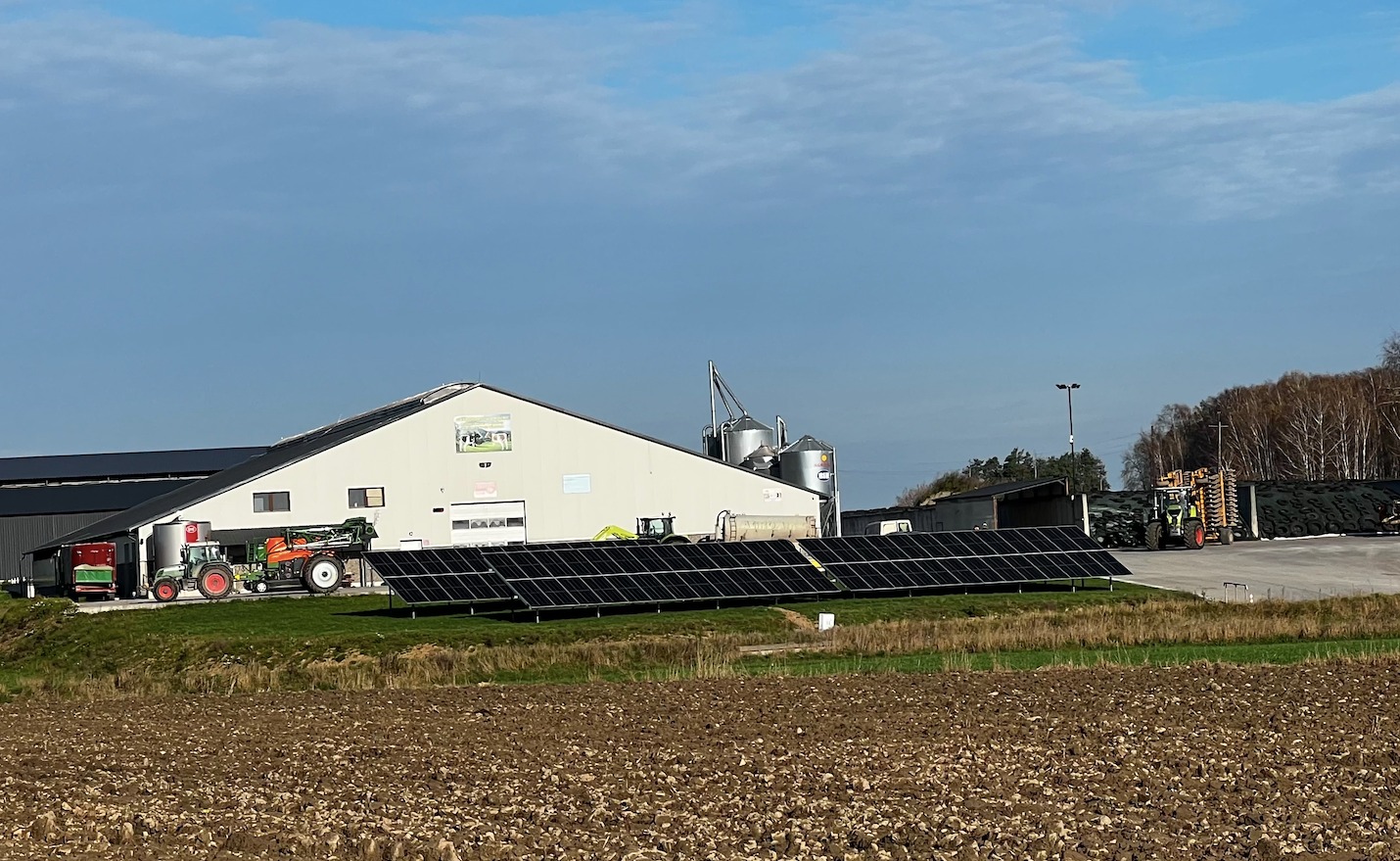 Panel słoneczny przed budynkiem gospodarczym z ciągnikami rolniczymi w tle, widok na instalację w pochmurny dzień.