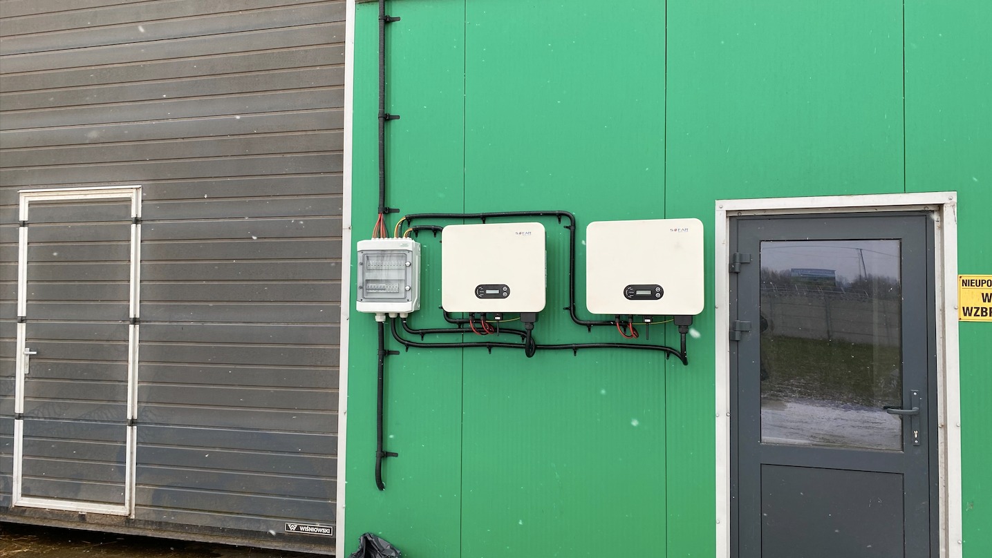 Dwa falowniki oraz skrzynka elektryczna zamontowane na zielonej ścianie kontenera, obok szary kontener z drzwiami.