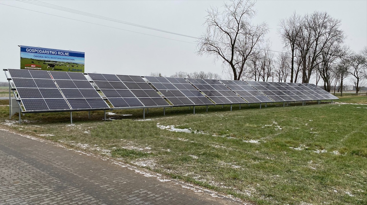 Rząd paneli słonecznych na metalowych stelażach, zainstalowany na terenie gospodarstwa rolnego, widoczny billboard reklamowy obory wolnostanowiskowej w tle, pochmurny dzień.