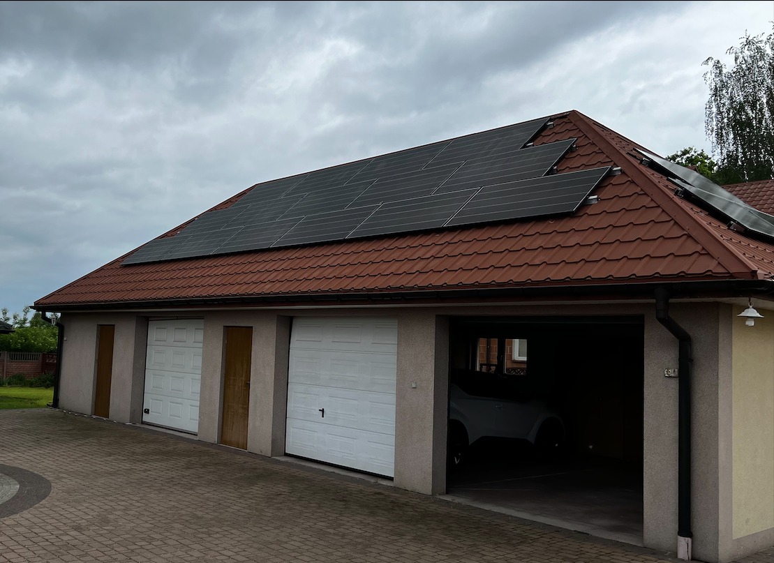 Panele słoneczne na dachu garażu z trzema bramami garażowymi, widoczne auto w jednym z garaży, pochmurne niebo