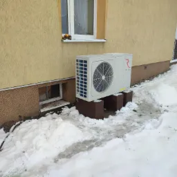 Biała pompa ciepła marki Rotenso, zamontowana na ceglanych podstawach przy otynkowanym budynku w zimowej scenerii, z narzędziami leżącymi na parapecie okna powyżej.