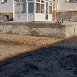 Przygotowanie fundament&oacute;w pod taras z białych bloczk&oacute;w, widoczna osoba z wężem ogrodowym, ziemia, kamienie i fragment elewacji budynku z oknami.