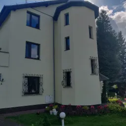 Jasna elewacja domu z wieżyczką, ozdobne kraty w oknach, widok z poziomu trawnika z kwiatami.