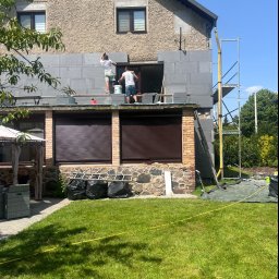 EDE GROUP SPÓŁKA Z OGRANICZONĄ ODPOWIEDZIALNOŚCIĄ - Dwóch pracowników ociepla elewację domu jednorodzinnego szarym styropianem, widoczne rusztowanie, zabezpieczona folią trawa i worki na odpady.