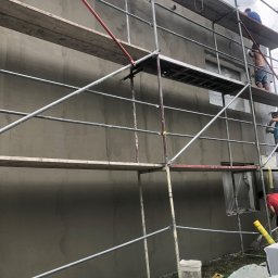 EDE GROUP SPÓŁKA Z OGRANICZONĄ ODPOWIEDZIALNOŚCIĄ - Ściana budynku w trakcie nakładania tynku zewnętrznego, widoczne rusztowania, pracownicy w trakcie pracy, wiadro z zaprawą i narzędzia.