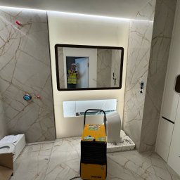 EDE GROUP SPÓŁKA Z OGRANICZONĄ ODPOWIEDZIALNOŚCIĄ - Wykończona łazienka w Słubicach z marmurowymi płytkami i podświetlanym lustrem. Widoczny osuszacz powietrza i elementy armatury w trakcie montażu.