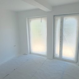 EDE GROUP SPÓŁKA Z OGRANICZONĄ ODPOWIEDZIALNOŚCIĄ - Białe, świeżo pomalowane wnętrze pokoju z dwoma oknami zabezpieczonymi folią malarską i zakrytym podłożem, widoczne gniazdka elektryczne.