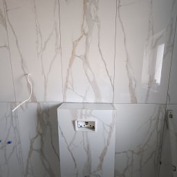 EDE GROUP SPÓŁKA Z OGRANICZONĄ ODPOWIEDZIALNOŚCIĄ - Łazienka w trakcie wykańczania: białe płytki imitujące marmur na ścianach, zabudowa stelaża WC, widoczne elementy instalacji. Poziomica przy ścianie.